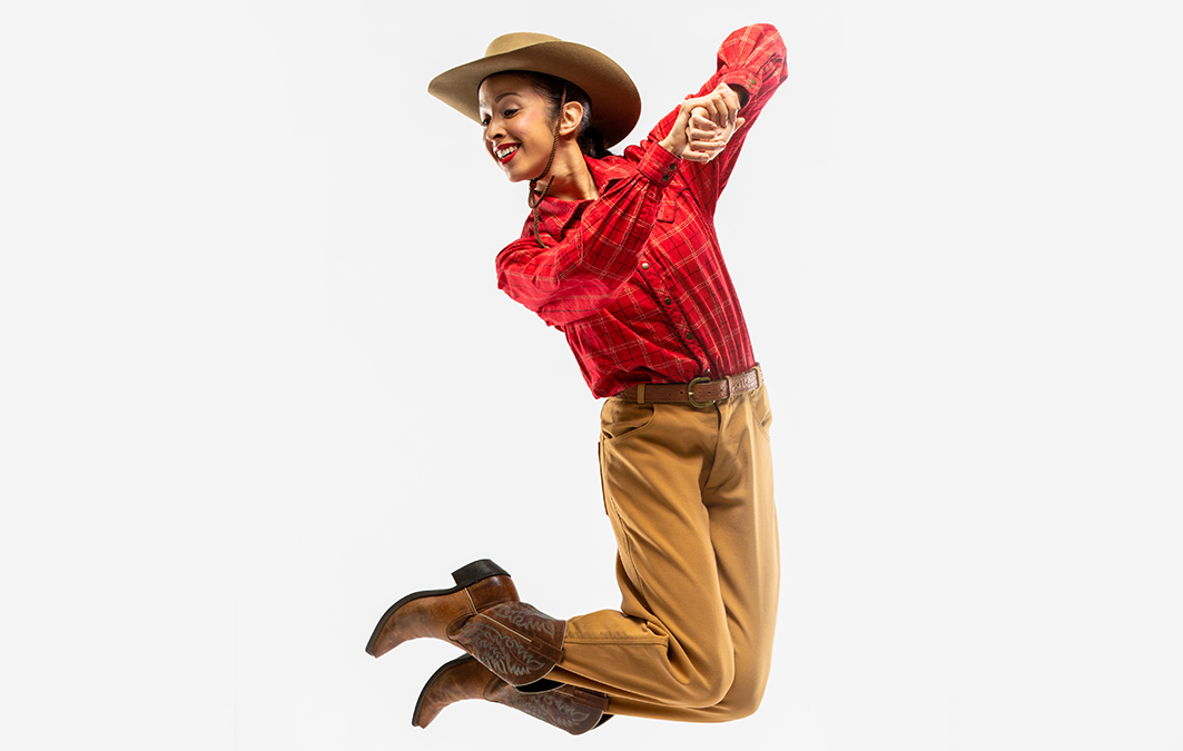 Jenna Rae Herrera jumps cheerfully in front of a white background, knees bent and clasping her hands in front of her chest with elbows bent to the sides. She wears brown cowboy boots, tan slacks, a bright red button down top and a tan cowboy hat.