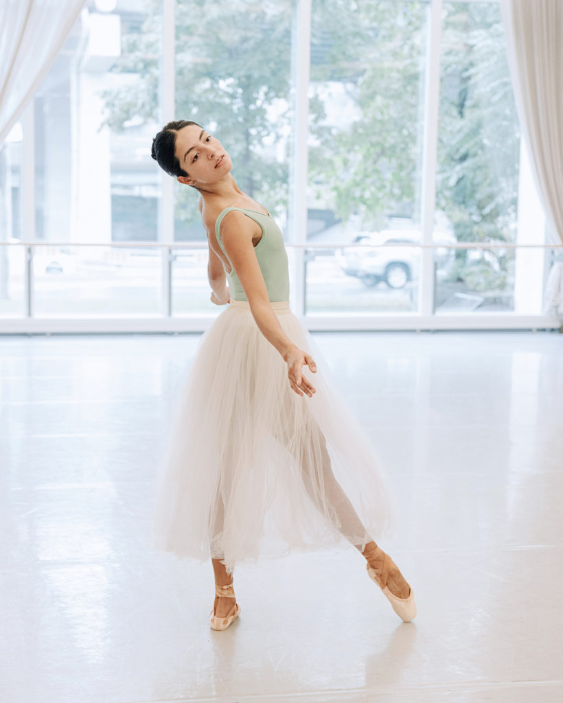 Selen Guerrero-Trujillo stands in profile with her right foot in tendu devant and her arms in demi-second, and leans her upper body back, looking over her right shoulder towards the front of the studio. She wears a mint green leotard, a long white tutu, beige tights and pink pointe shoes.