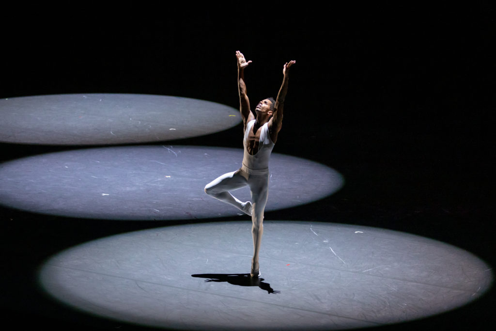 Jonathan Batista dances onstage during a performance, doing a passé relevé with his right leg up and his arms stretched above his head. He wears a white unitard and looks up toward the ceiling, bathed in a spotlight on an otherwise darkened stage.