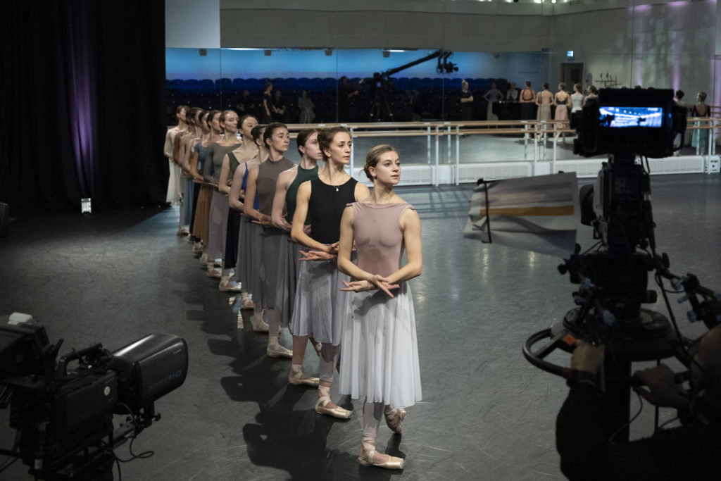 A line of female dancers pose in B plus, arms crossed low, at the side of the studio as camera operators watch from the front of the room. The dancers wear various colors of leotards and long rehearsal skirts.