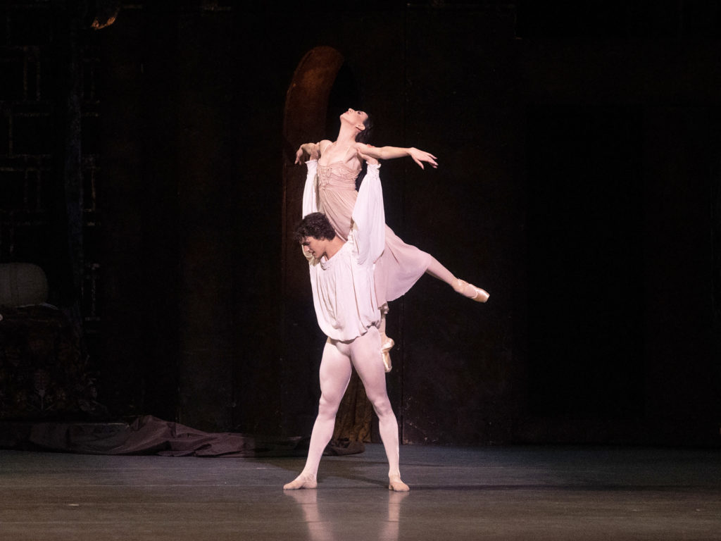 Daniel Camargo, wearing a loosely fitting white peasant blouse, white tights and white ballet sippers, stands in a wide stances and holds Isabella Boylston over her head by her forearms. Boylston, in a pink nightgown, makes a small arabesque and looks up towards the ceiling.