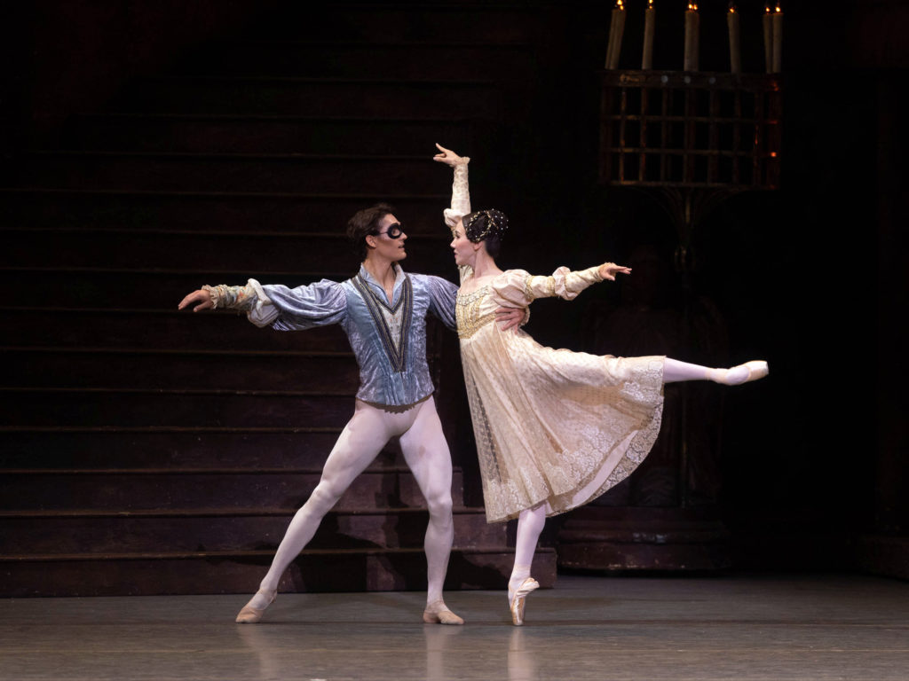 Daniel Camargo, costumed in a blue tunic, white tights and ballet slippers, and a black eye-mask, holds onto the waist of Hee Seo as she balances in arabesque on pointe. The dancers look at each other in the eyes, and Seo wears an off-white empire-waist gown, pink tights and pink pointe shoes.