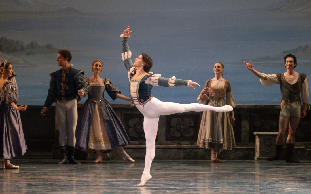 During a performance of "Swan Lake," Daniel Camargo poses in first arabesque with his left leg raised and his right arm curved above his head. He wears a green and white tunic, white tights and white ballet slippers. Behind him, dancers dressed in courtier costumes watch him perform.