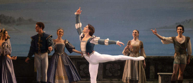 During a performance of "Swan Lake," Daniel Camargo poses in first arabesque with his left leg raised and his right arm curved above his head. He wears a green and white tunic, white tights and white ballet slippers. Behind him, dancers dressed in courtier costumes watch him perform.