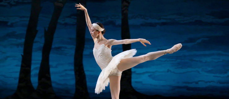 Yuriko Kajiya does a first arabesque with her left leg up during a performance of Swan Lake. She wears a white tutu and white-feathered headpiece, pink tights and pink pointe shoes. Her eyes are downcast, and she lifts her right arm up high, near her right ear, Behind her is a blue backdrop with the black silhouettes of trees.