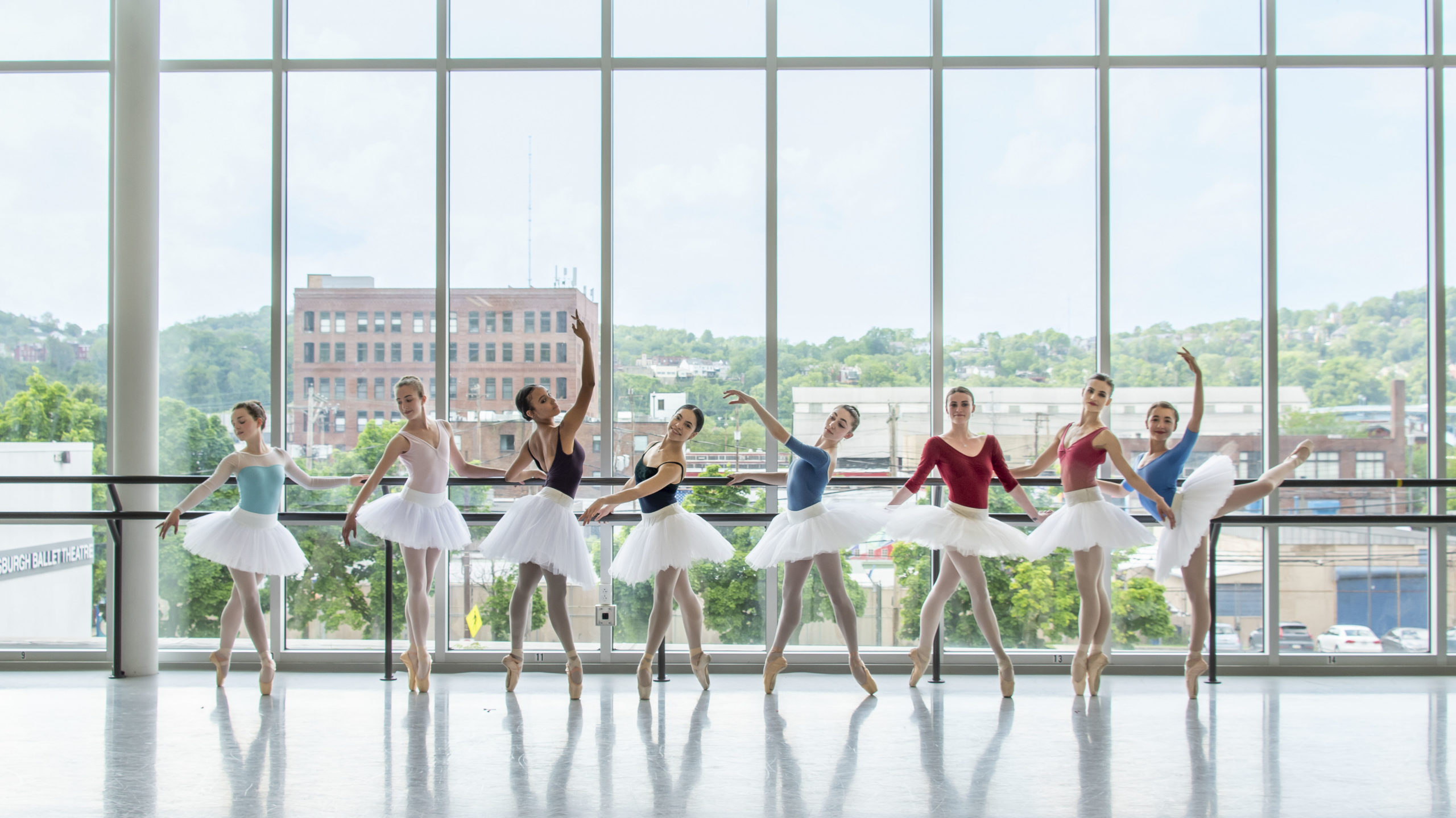 Eight young adult female ballet dancers in bright leotards, pointe shoes and white tutus pose in a line in front of a barre and a wall of windows.