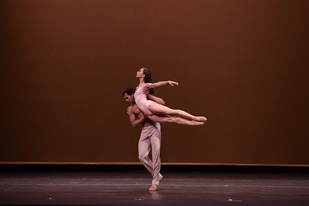 Denise Phillips and Joshuan Vazquez dance together onstage during a performance. Vazquez, shirtless and wearing loose tan pants, lifts Phillips in the air holding her under the left shoulder and right hip. Phillips, in a pink leotard and ballet shows, lengthens her legs behind her and arches her back, wrapping her right arm around Vazquez's shoulders and extending her left arm out to the side.