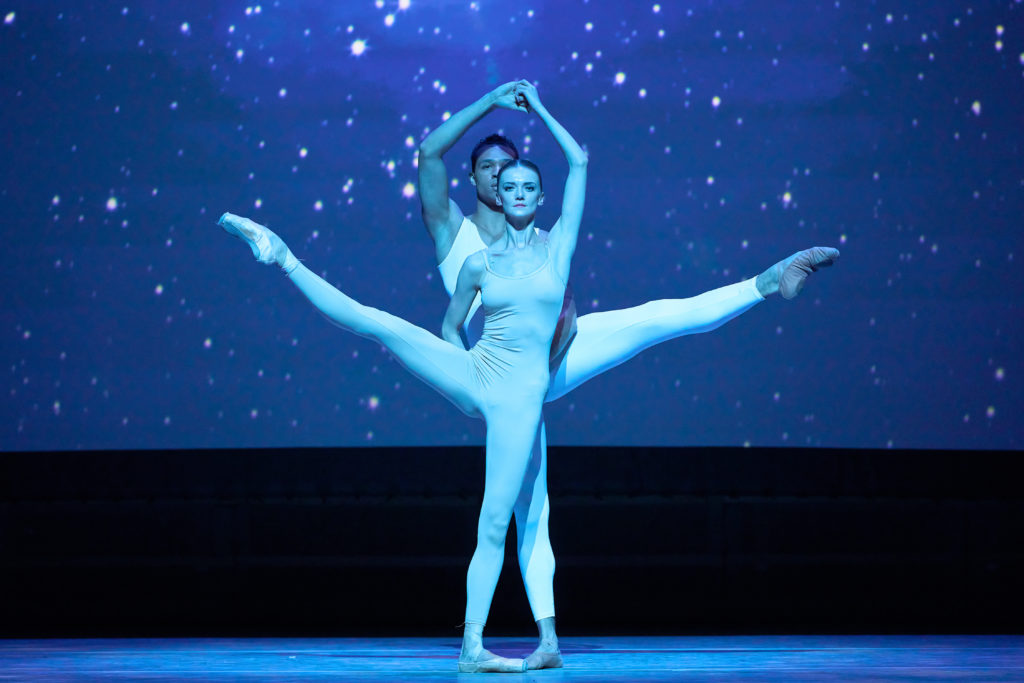 Andrea Laššáková and Adrian Blake Mitchell wear white unitards in front of a galaxy-like backdrop onstage. Lassakova stands in front of Black Mitchell and they mirror each other, one leg held a la seconde, opposite hands holding each others' above their heads and behind Lassakova's back.