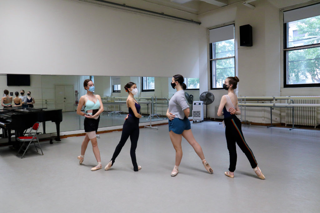 Four college-age female dancers rehearse in a studio. In pairs, they face in toward each other in a diagonal line, posing in tendu derrierre. They wrap their arms around their own waists and wear face masks, pointe shoes and various dancewear.