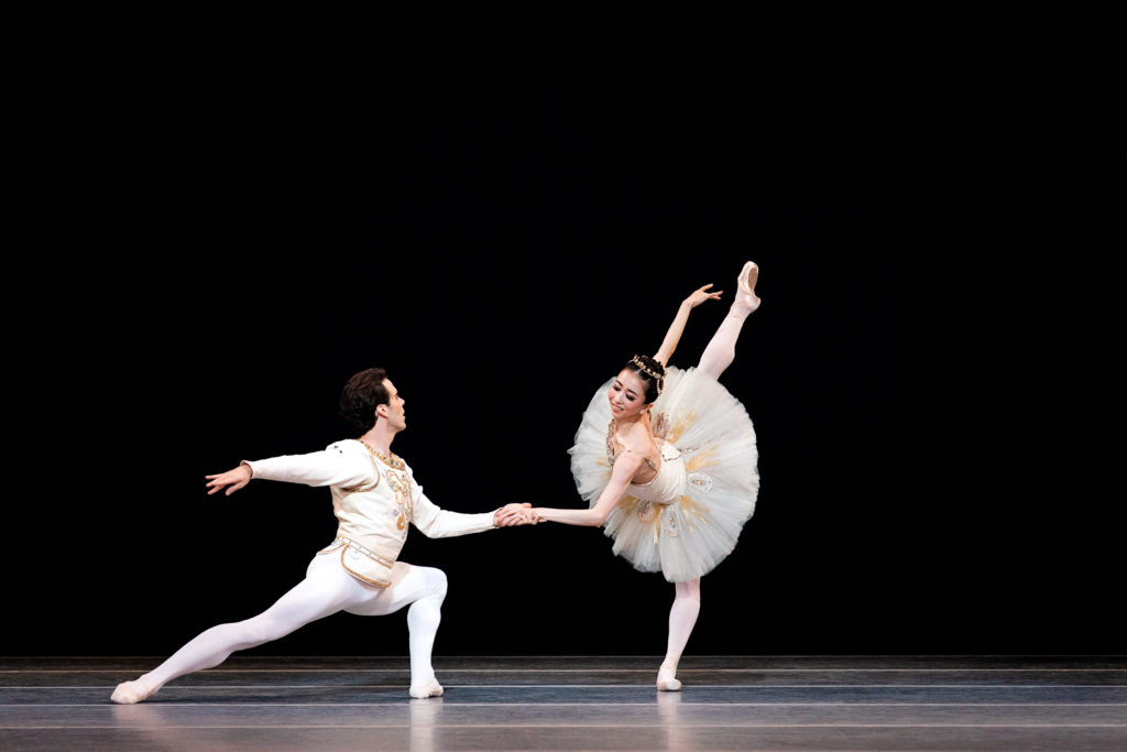 Connor Walsh lunges low, extending his right leg back, facing his partner, Yuriko Kajiya and holding her hadn as she performs a penché arabeque croisé on flat. She wears a white tutu with gold trim, a tiara, pink tights and pointe shoes, while he wears white tights and a white tunic with gold trim. They perform onstage in front of a black backdrop.