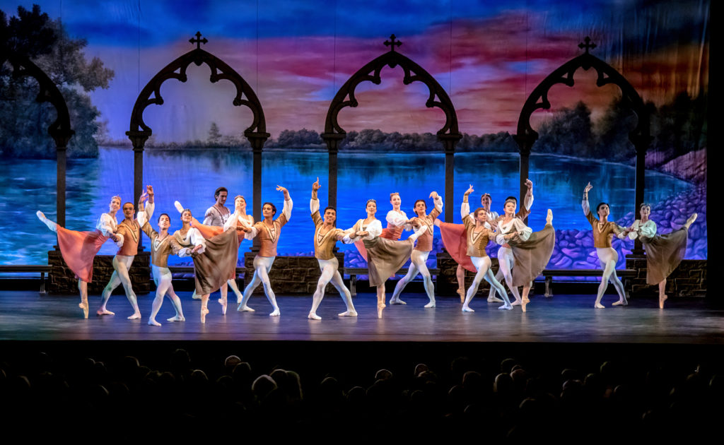A large group of male and female dancers poses together onstage during a performance. The men stand in a large fourth position and offer one hand, while a female dancer holds on to it and to his shoulder and balance in attitude derriere. Themen wear white tights and ballet slippers and tan tunics, while the ladies wear calf-length dresses in nuetral tones and with long, white puffed sleeves, pink tights and pointe shoes.