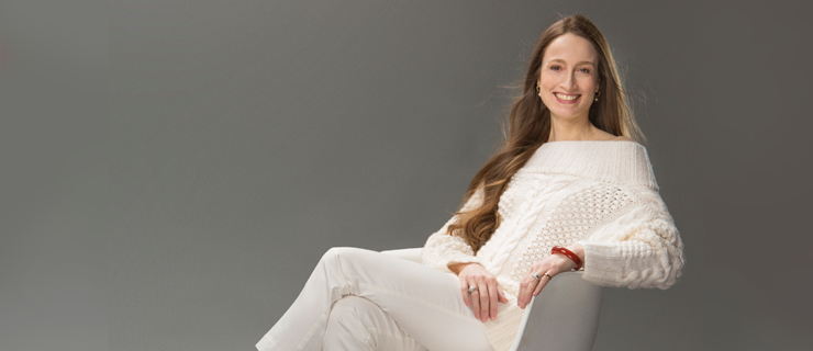 Julie Kent sits in a white chair, legs crossed and hands in her lap. She smiles at the camera and wears tan high heels, white slacks and a white sweater.