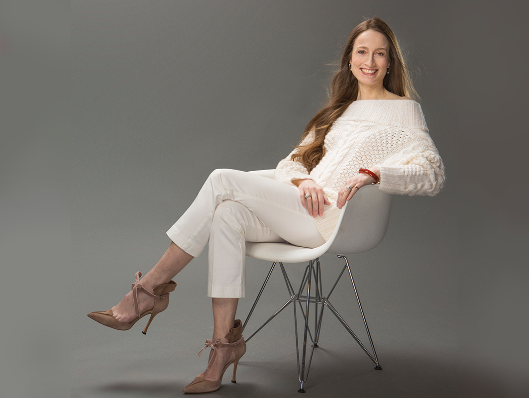 Julie Kent sits in a white chair, legs crossed and hands in her lap. She smiles at the camera and wears tan high heels, white slacks and a white sweater.
