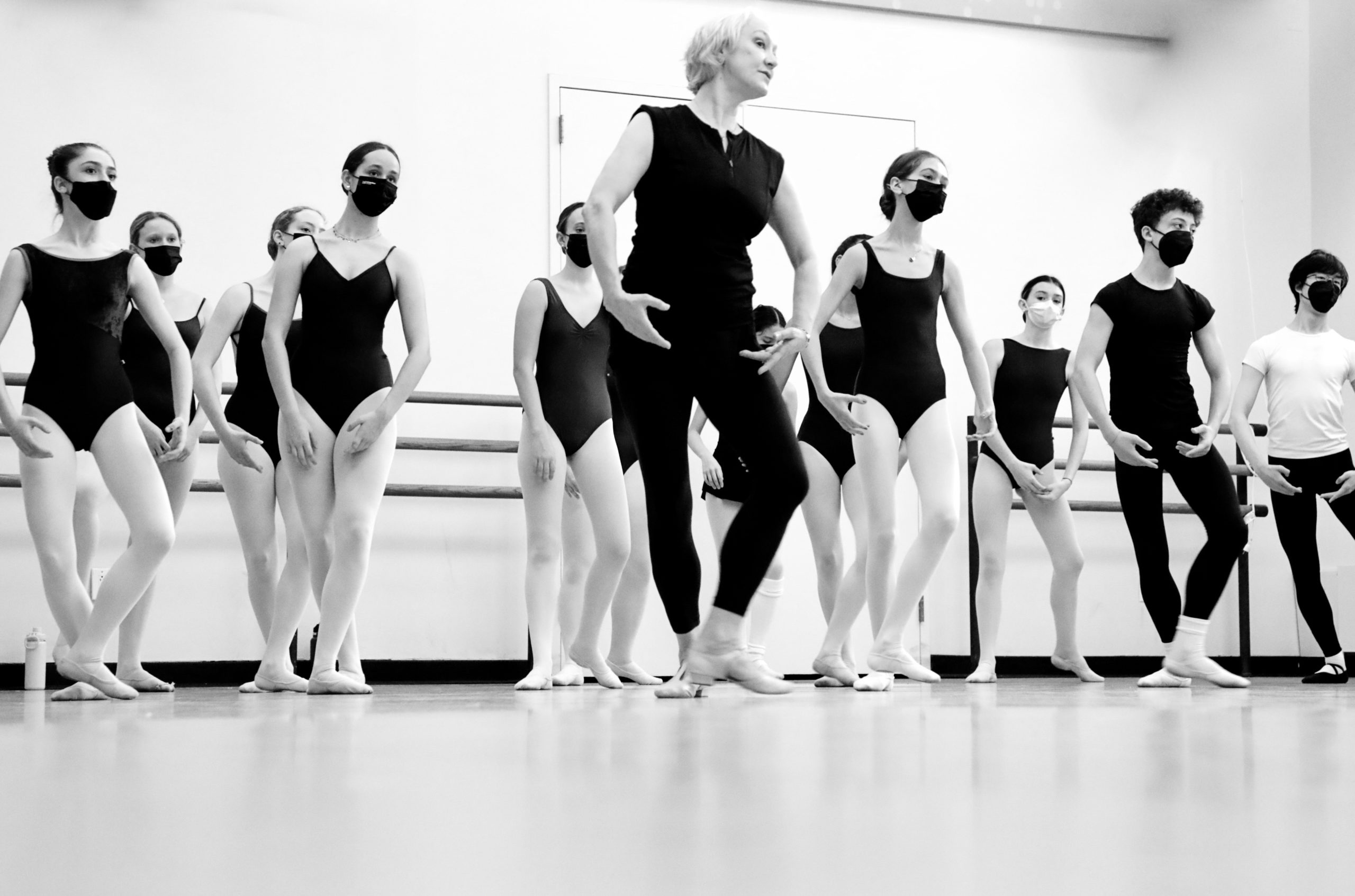 In this black and white photo, Deborah Wingert stands at the front of a ballet class and demonstrates a step moving through fifth position with her arms en bas. Behind her, a large class of students wearing dance clothing, ballet slippers and face masks copy her movements.