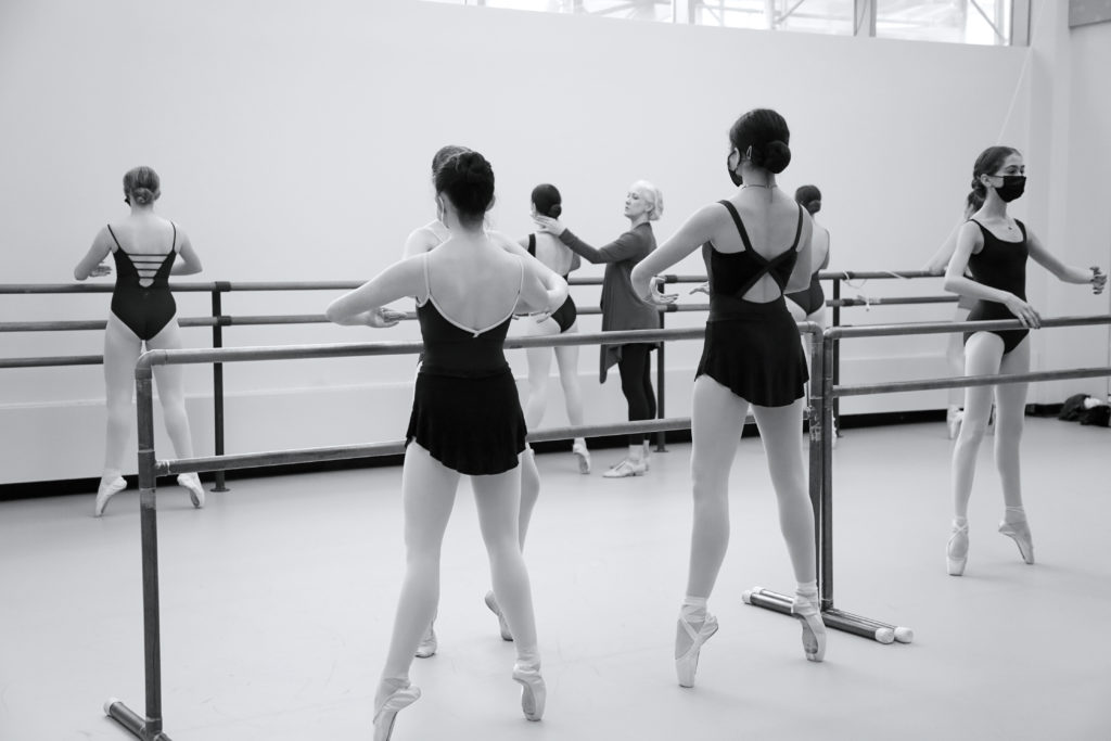 In this black and white photo, a group of ballet students practice balancing in first position relevé on pointe, facing the barre. They wear black leotards, short ballet skirts, tights and pointe shoes. Deborah Wingert adjusts the head of one dancer. She wears a long sweater, black tights and teaching shoes.