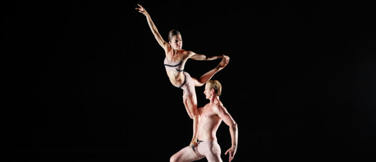 A woman ballerina balances by standing on one leg on a male dancer's thigh, who is positioned in a lunge.