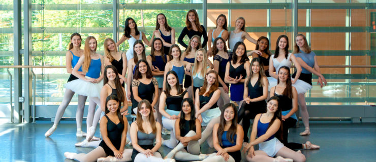A large group of college-age, female dancers in various leotards, tights, skirts and pointe shoes pose in a large formation in front of floor-to-ceiling windows in a dance studio.