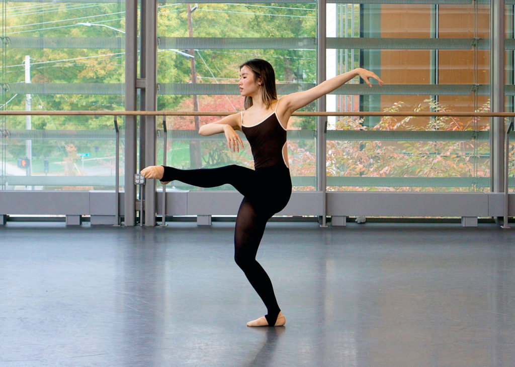 Alyssa Shi poses in attitude devant with her right leg in plié and her left leg raised. She wears a black leotard, black tights and ballet slippers. She poses in front of a large window in a dance studio.