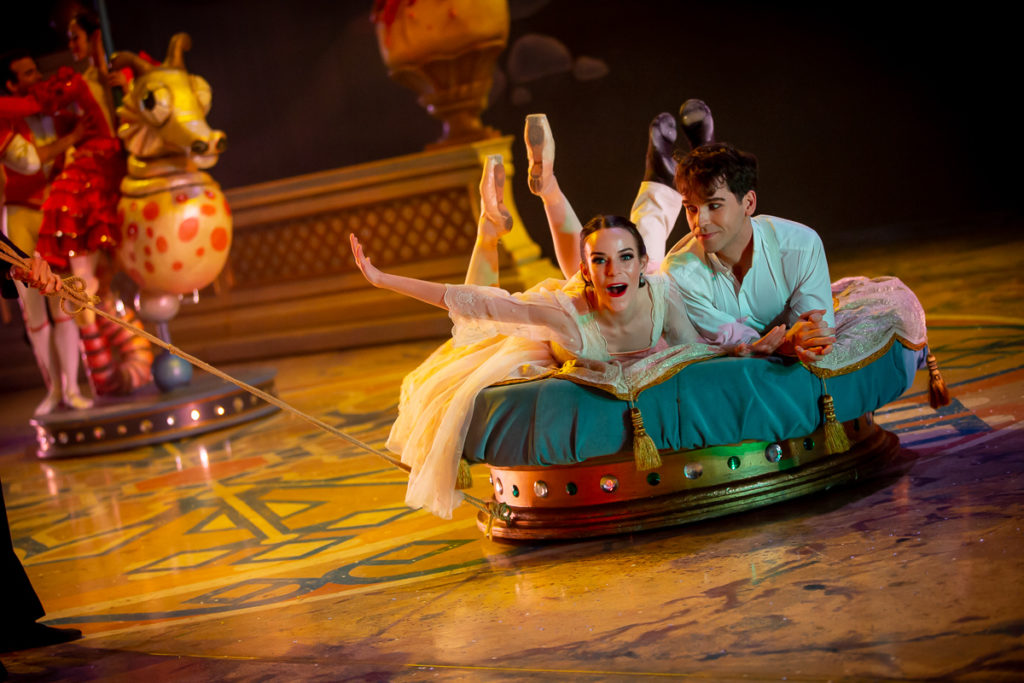 Elizabeth Harrison and Marko Micov lay propped on their stomachs on a large, blue circular settee, kicking their legs up behind them. Harrison reaches her right arm out to the side and wears a gleeful expression, and wears a filmy pink dress and pointe shoes. To her left, Micov wears a white shirt and pants and black ballet slippers, and looks at Harrison with a small smile.