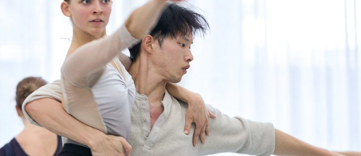 Shown hips-up in a brightly lit studio, Genevieve Penn Nabity and Kota Sato dance together. Sato holds Nabity around her back as she reaches forward intently in an arabesque.