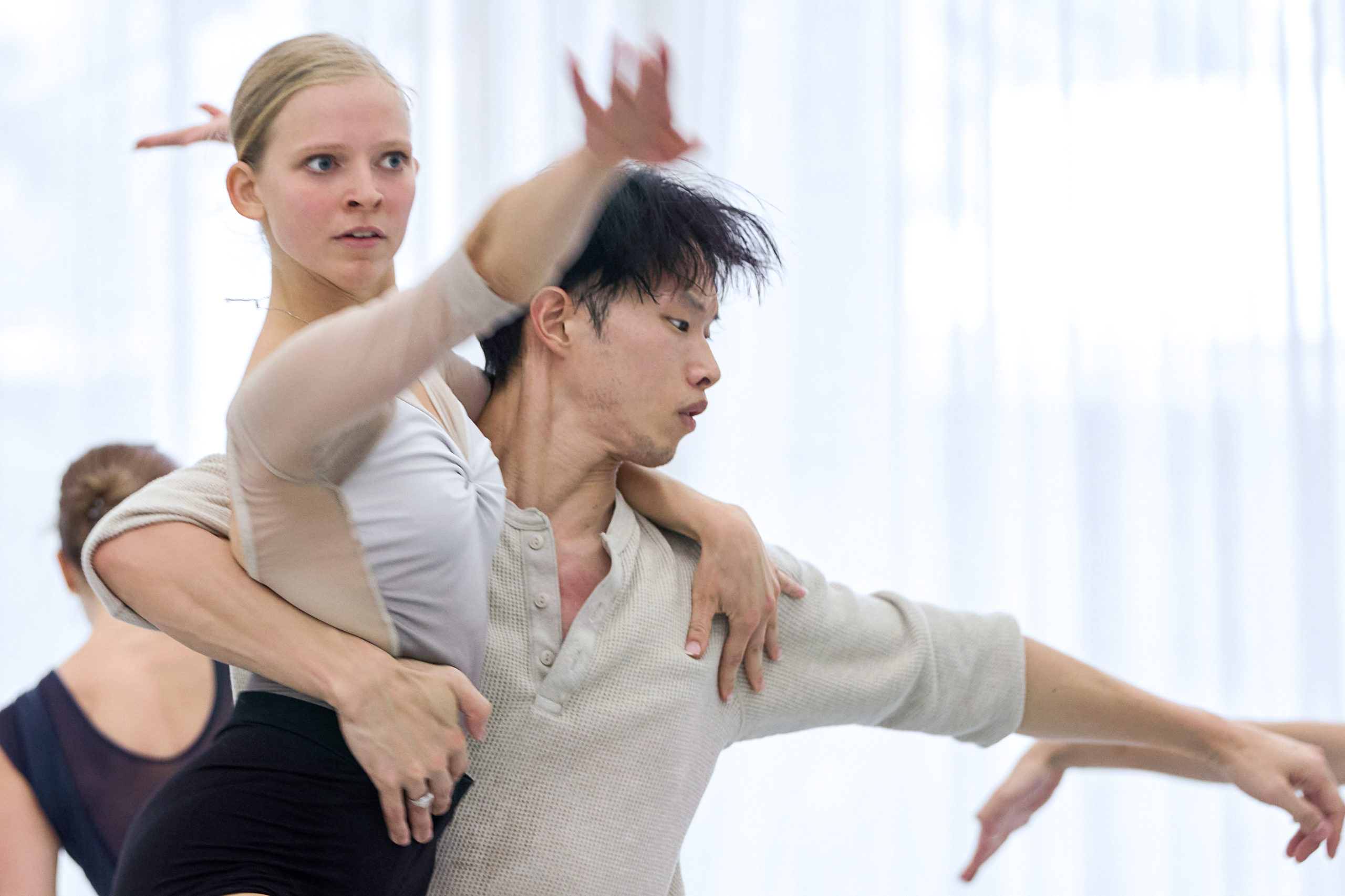 Shown hips-up in a brightly lit studio, Genevieve Penn Nabity and Kota Sato dance together. Sato holds Nabity around her back as she reaches forward intently in an arabesque.