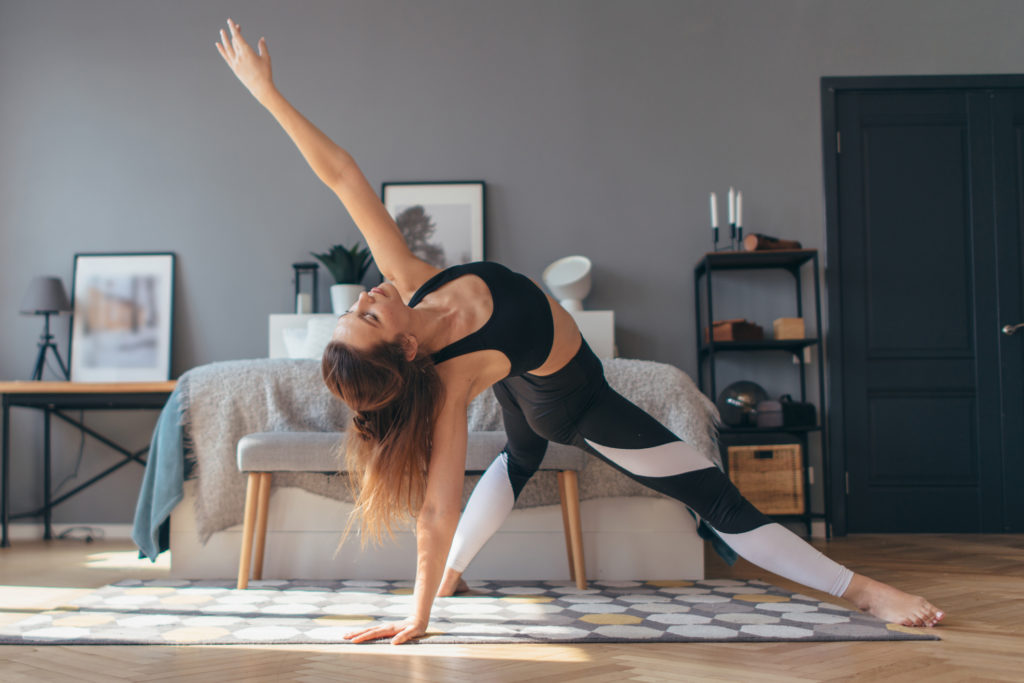A woman in athletic clothing and a ponytail does a "wild thing" yoga pose on a spotted yoga mat in a cozy bedroom.
