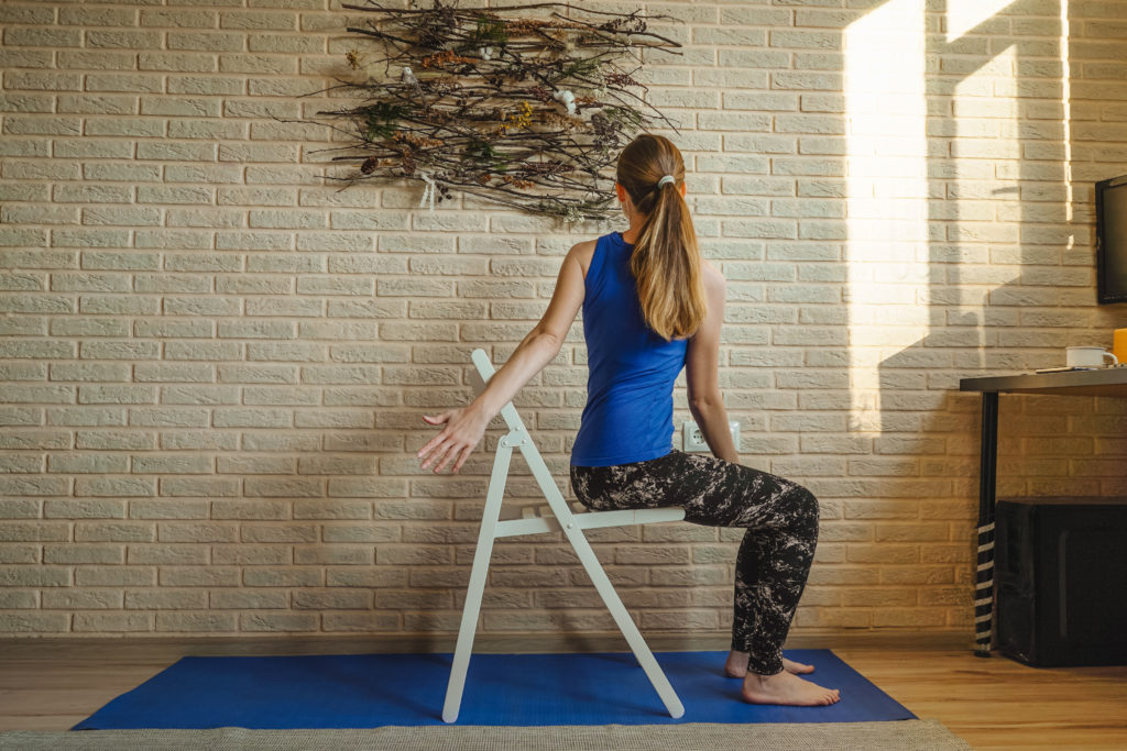 A woman in a blue top and black marble-print leggings twists her back sitting on a folding chair on top of a blue yoga mat. A yellow brick wall and natural-looking decoration are in the background.