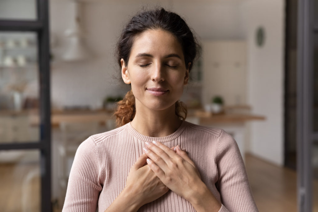 Close up of calm young Caucasian woman hold hands on heart while meditating.