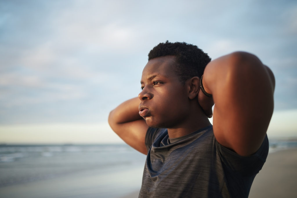 Cloesup of a Black young man with hands behind his head. He is standing on a beach looking stressed.