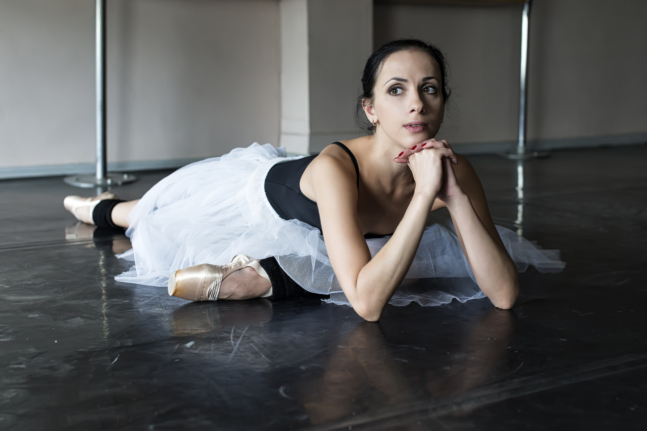 A ballerina in a black camisole leotard and white practice romantic tutu, in pointe shoes, stretches on the floor of a ballet studio with a pondering look on her face. She rests her chin on her hands, which are folded together.