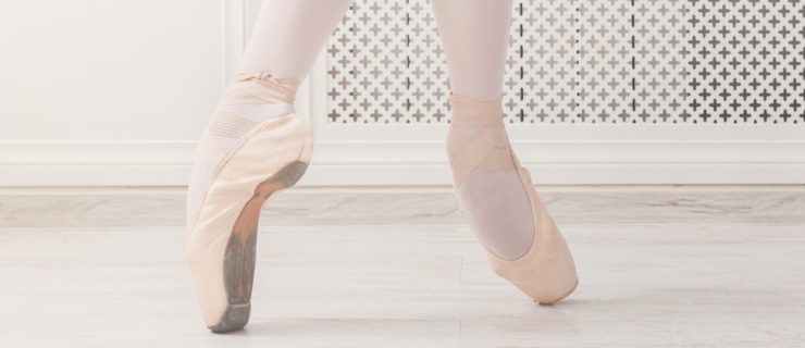 A ballerina is shown from the knees down on pointe, wearing pink tights, on a white wooden floor..