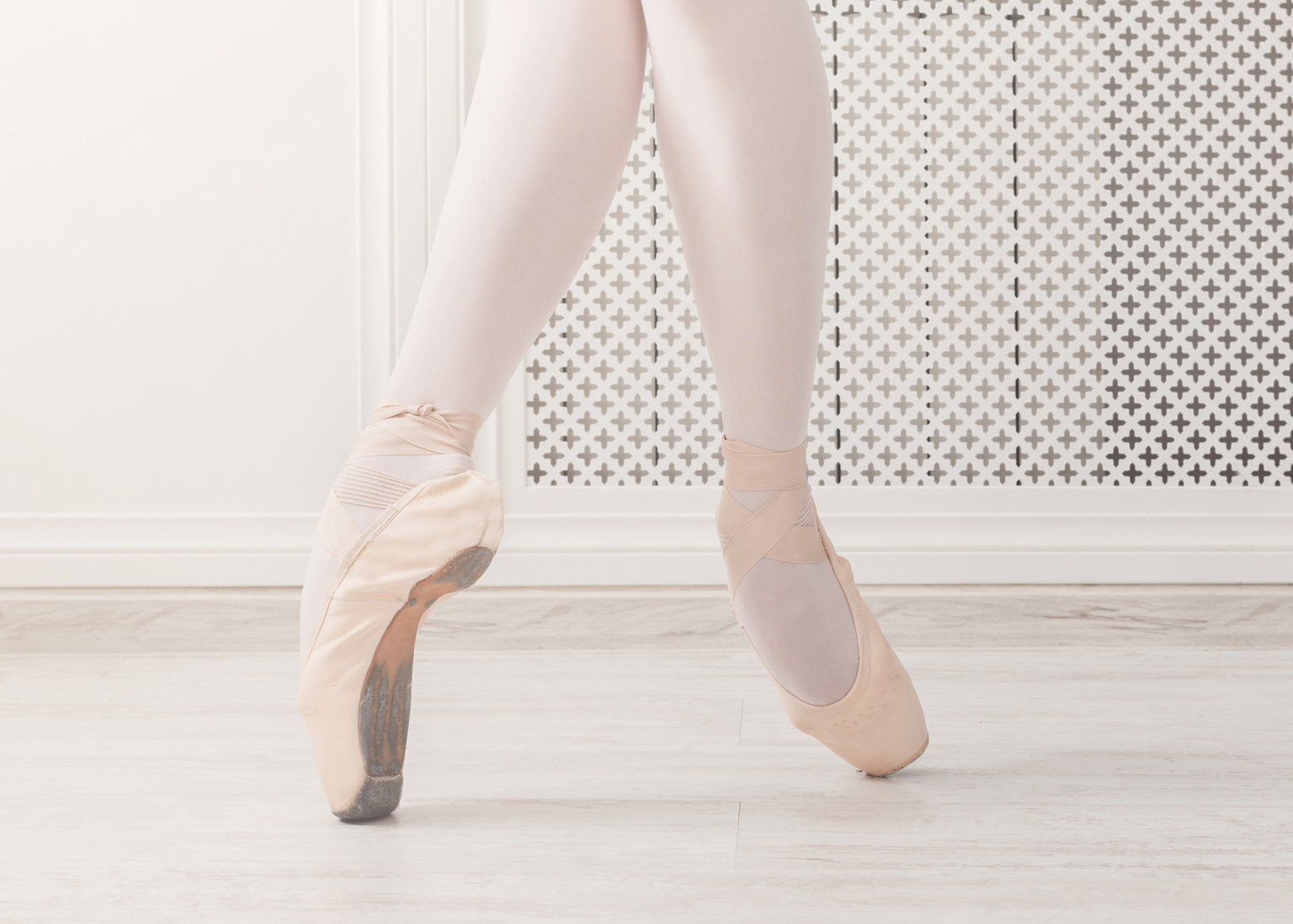 A ballerina is shown from the knees down on pointe, wearing pink tights, on a white wooden floor..