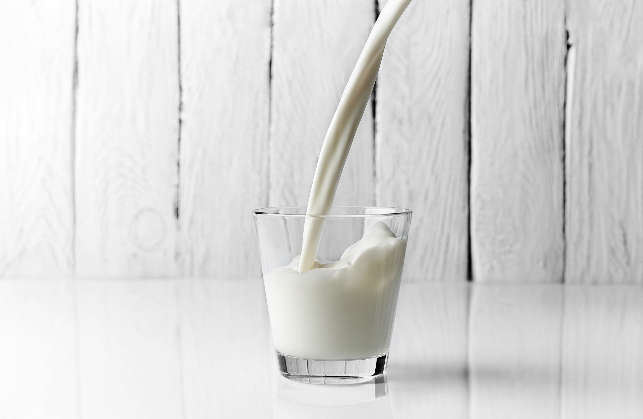 Milk being poured into a glass in front of a white wooden background.