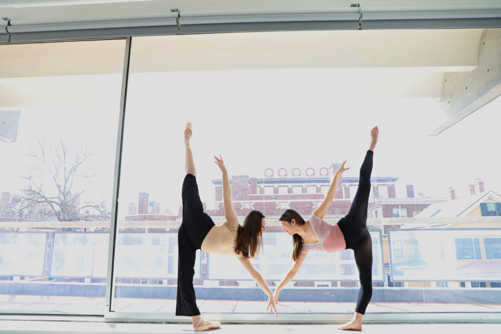 Nina Montalbano and Sara Komatsu face in to each other and do a penché in front of a large window in a dance studio. Montalbano wears pointe shoes, loose black pants and a peach leotard. Komatsu wears a pink leotard and black tights, and is barefoot. They both wear their long hair down.