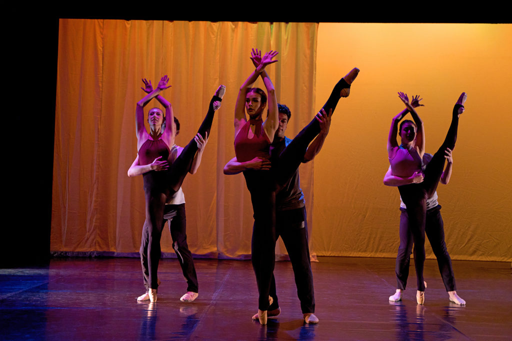 A trio of male/female partners perform onstage, making a triangle shape with one couple center stage and the others on either side. The women lift their left leg into a high développé a la seconde and cross their wrists over their heads; they wear halter top leotards, stirupped black tights and pointe shoes. The men, wearing black pants, ballet slippers and T-shirts, stand behind their partner, holding their waist with their right arm and the women's lifted leg with their left hand.