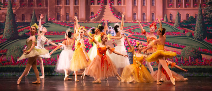A group of young female dancers pose in a tableau onstage in front of a backdrop showing a grand palace with rose gardens. They wear tuts in various colors and lengths. Cassidy Jarvis poses in the middle in a tendu derriere in plié, wearing an orange Romantic-length tutu with floral details on the bodice and a floral headpiece.