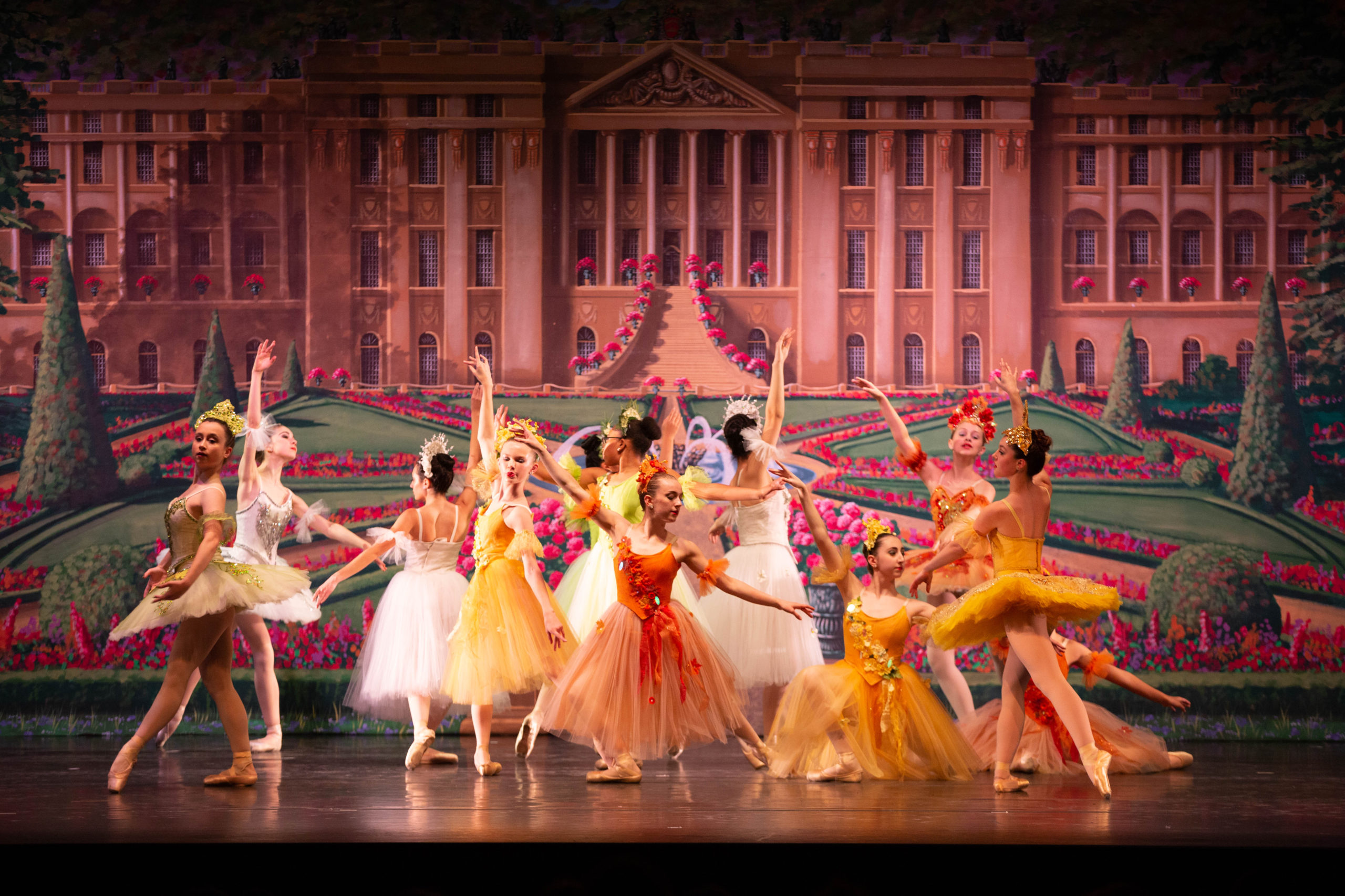 A group of young female dancers pose in a tableau onstage in front of a backdrop showing a grand palace with rose gardens. They wear tuts in various colors and lengths. Cassidy Jarvis poses in the middle in a tendu derriere in plié, wearing an orange Romantic-length tutu with floral details on the bodice and a floral headpiece.