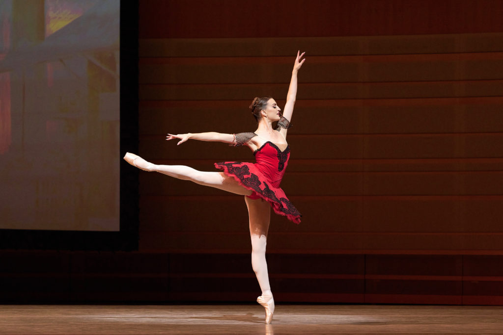 Joy Womack wears a bright red tutu with black lace embellishment, pink tights and pointe shoes as she arabesques energetically onstage. She performs the role of Kitri in "Don Quixote."