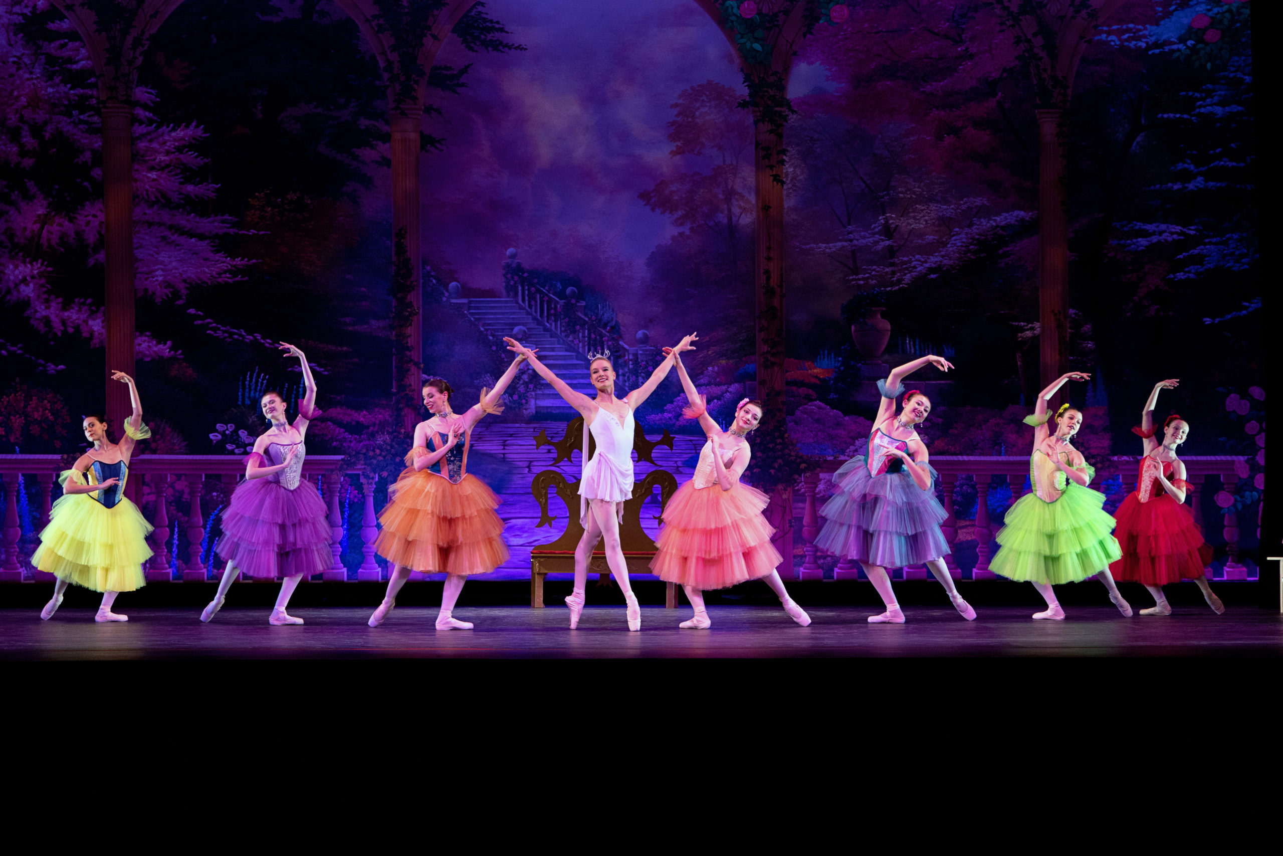 Lauren Strenoos, in a short, white dance dress, pink tights and pointe shoes, poses center stage in a fourth position on pointe, her arms in a wide V-shape. A line of corps women are on either side of her in a tendu derriere, with their inside leg in plié. They wear brightly colored, Spanish-style long tutus and pointe shoes. The two closest to Strenoos hold on to her wrists.