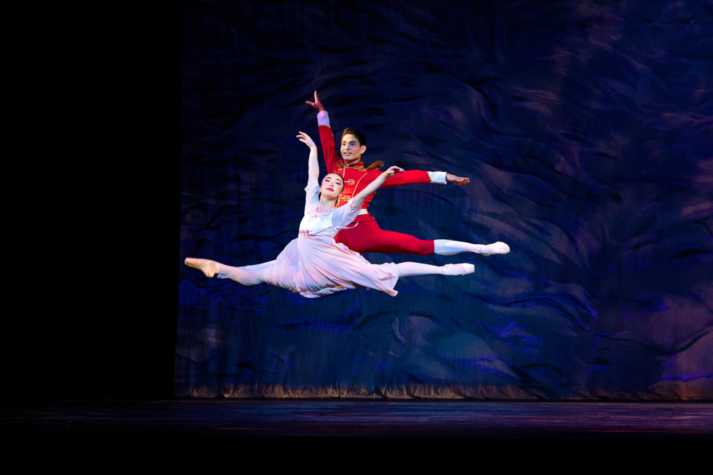 Marla Aleyda and Bruno Palheta both do a large saut de chat together onstage during a performance of The Nutcracker. Palheda, in a red soldier costume with white tights and ballet slippers, jumps directly behind Aleyeda, who wears a pink filmy dress, pink tights and pointe shoes. They look out towards the audience and dance in front of a dark backdrop.
