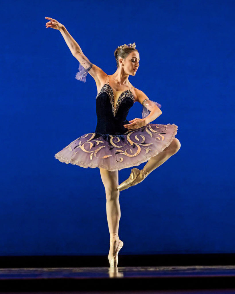 Katherine Barkman wears a black ombre tutu that fades to white at the skirt edges, embellished with gold swirls. She stands en pointe in retire, arms in 3rd, as she performs a variation onstage in front of a dark blue backdrop.