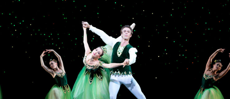 Tracia Albertson and Rainer Krenstetter perform onstage in front of a black backdrop with green sparkles. Krenstetter holds Albertson by both wrists as she leans forward into a penché arabesque on pointe with her right leg back. She wears a green Romantic tutu with emerald rhinestones on the waist, neckline and tiara. Krenstetter wears a green velvet top, white tights and white ballet slippers. Tow female dancers in similar green tutus kneel on the ground behind them with their arms in high fifth position.