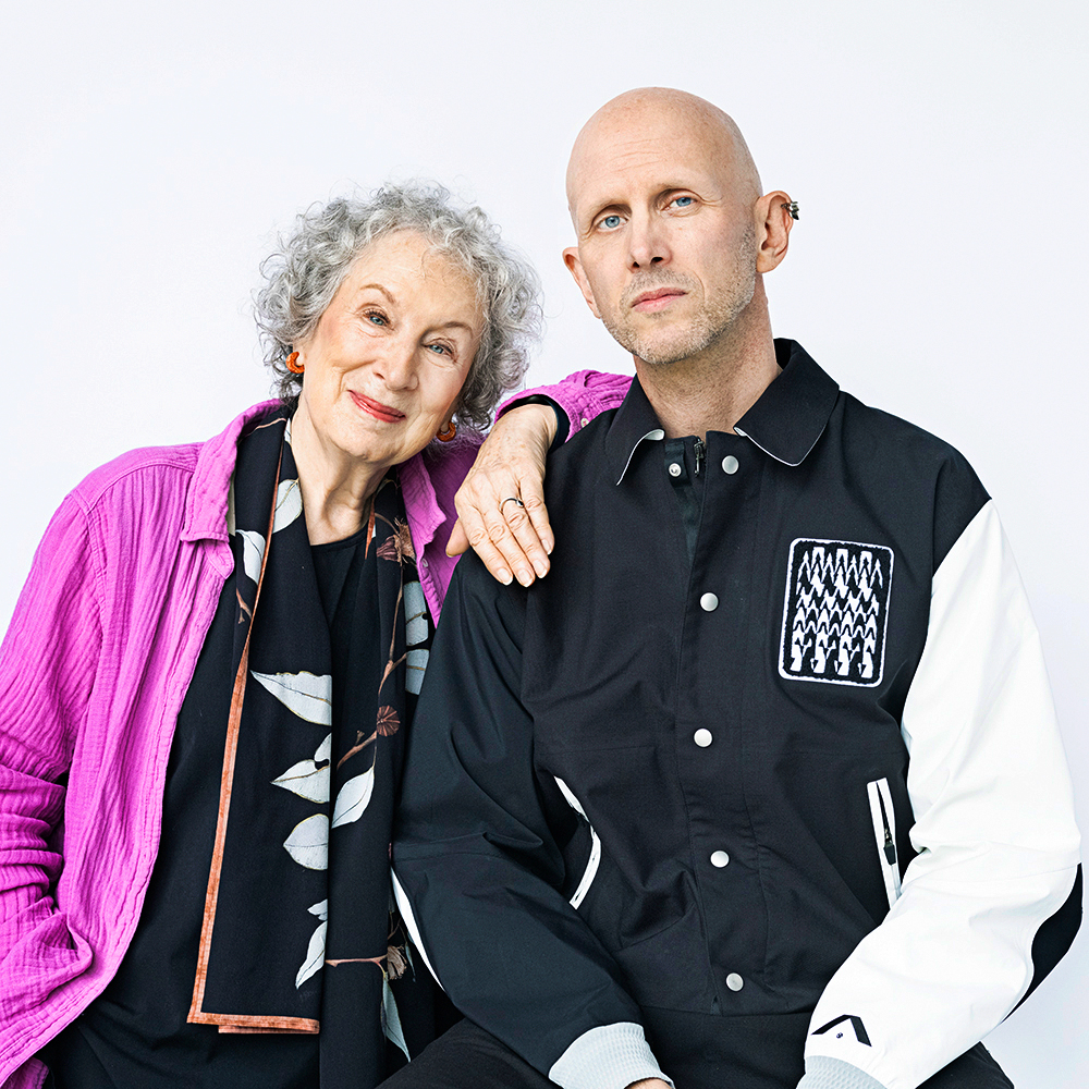 Margaret Atwood and Wayne McGregor sit together for a portrait photo in front of a white backdrop. Atwood has curly silver hair and wears a black shirt, pink button down and black and white scarf. She rests her left arm on McGregor's shoulder. McGregor, bald, wears a black and white modern button-down.