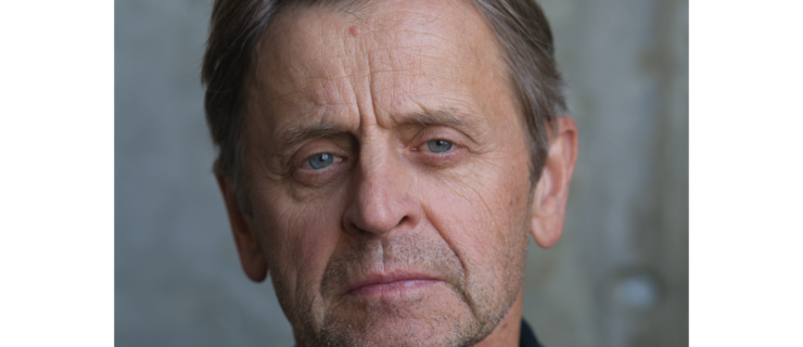 Mikhail Baryshnikov is photographed from the neck up. He wears a black shirt and poses in front of a gray wall. He looks directly at the camera with a calm expression on his face.
