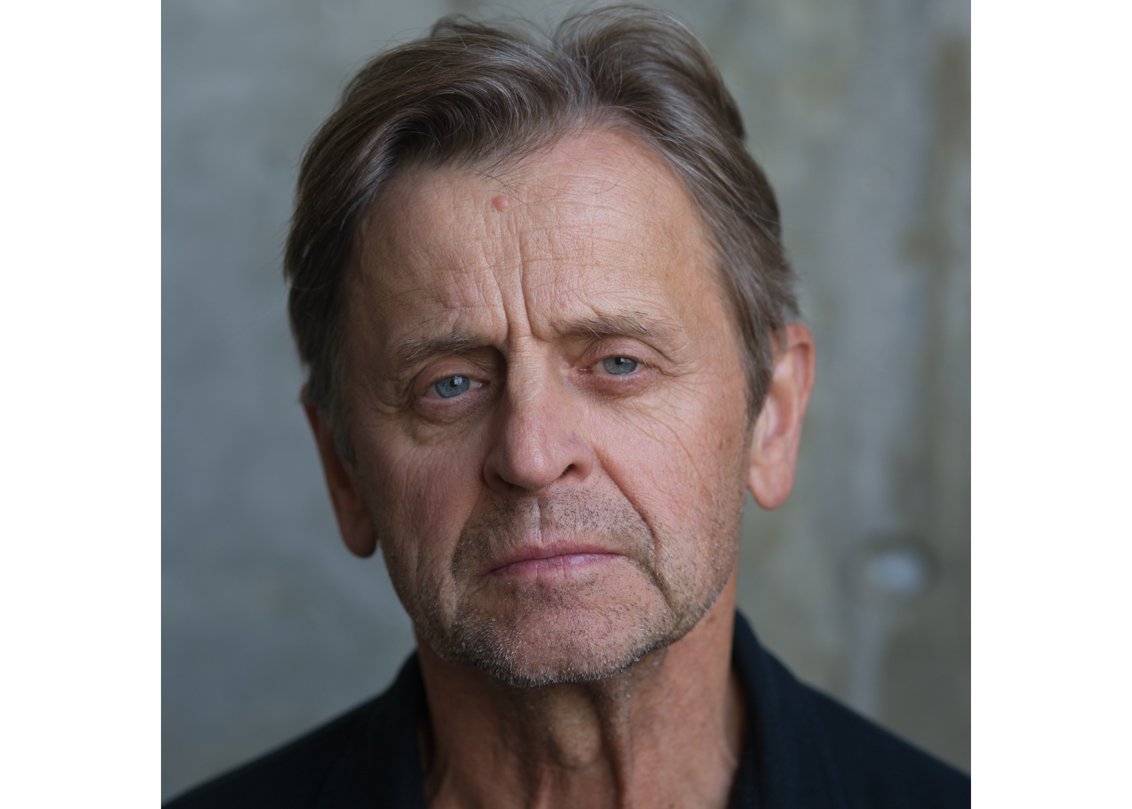 Mikhail Baryshnikov is photographed from the neck up. He wears a black shirt and poses in front of a gray wall. He looks directly at the camera with a calm expression on his face.