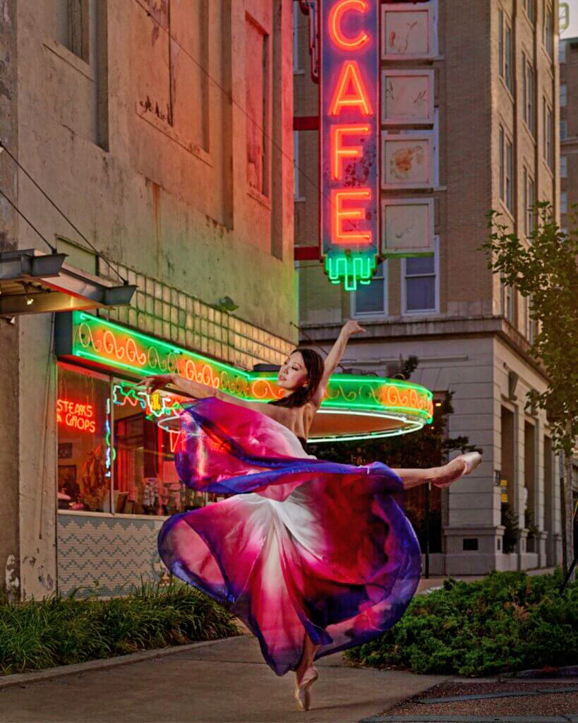 Misa Kuranaga does a sweeping attitude balance in a colorful, flowy long pink, white and purple dress in front of a city scape with neon lights.
