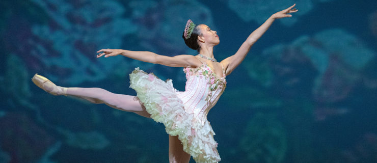 During a performance of "The Nutcracker," Nikisha Fogo does a first arabesque on pointe with her right leg up. She wears a light pink tutu with green trim and small flowers along the bodice, and a pink and green tiara, pink tights and pointe shoes.