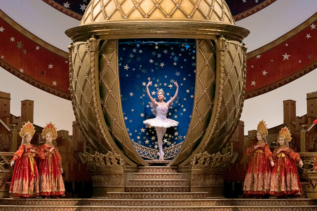 Tina Pereira stands in sus-sous on pointe inside a gilded, golden set. Behind her is a blue background covered in stars. She wears a pale pink tutu, gold tiara, pink tights and pointe shoes.