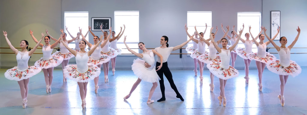 Four lines of young women in white and pink tutus on pointe. Between the lines a couple poses.