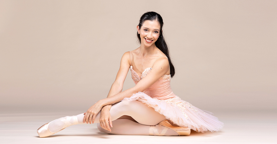 Macarena Giménez, wearing a pink tutu, tights and pointe shoes, sits on the ground and tucks her right foot underneath her. She extends her left leg forward, bending her knee and pointing her foot, and rests her crossed wrists over her calf. She wears her long hair down and looks towards the camera and smiles.